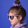 kleines Mädchen mit lila Sonnenbrille in Herzform steht seitlich im Profil, mit einem lila geblümten Scrunchie und hell lila Pullunder vor blauem Hintergrund.