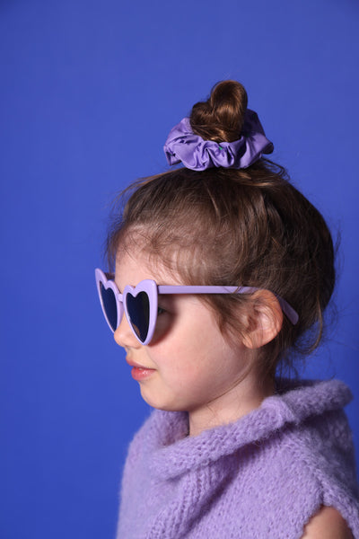 kleines Mädchen mit lila Sonnenbrille in Herzform steht seitlich im Profil, mit einem lila geblümten Scrunchie und hell lila Pullunder vor blauem Hintergrund.
