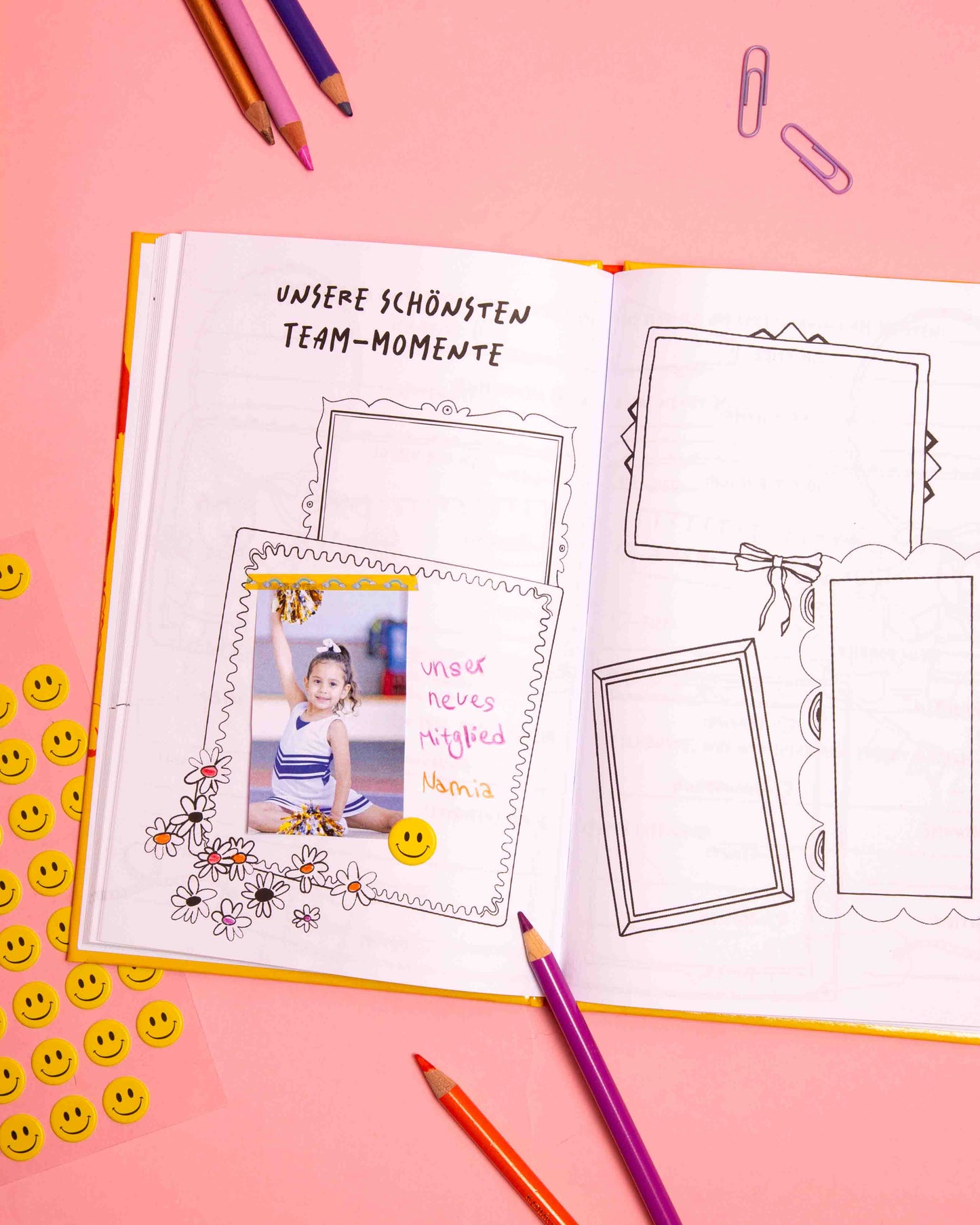 Cheerleading Freundebuch Beispielseite – Unsere schönsten Team-Momente. Aufgeklapptes Buch liegt auf rosafarbenem Hintergrund. Es liegen bunte Stifte, Klammern und Sticker drumherum.