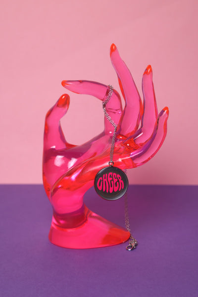 Cheerleader Medaillon in 925er Silber Kette und Edelstahl Anhänger, der mit dem Schriftzug Cheer versehen ist und Epoxidharz beinhaltet hängt an einer Pinken Plexiglashand auf einem lila Hintergrund.