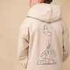 Junge steht mit dem Rücken zum Betrachter. Er trägt eine graue Capi, einen beigen Pullover mit einem großen Backprint mit einer One Line Zeichnung eines Cheerleading Groupstunts und der Aufschrift Cheerleader vor beigem Hintergrund. 