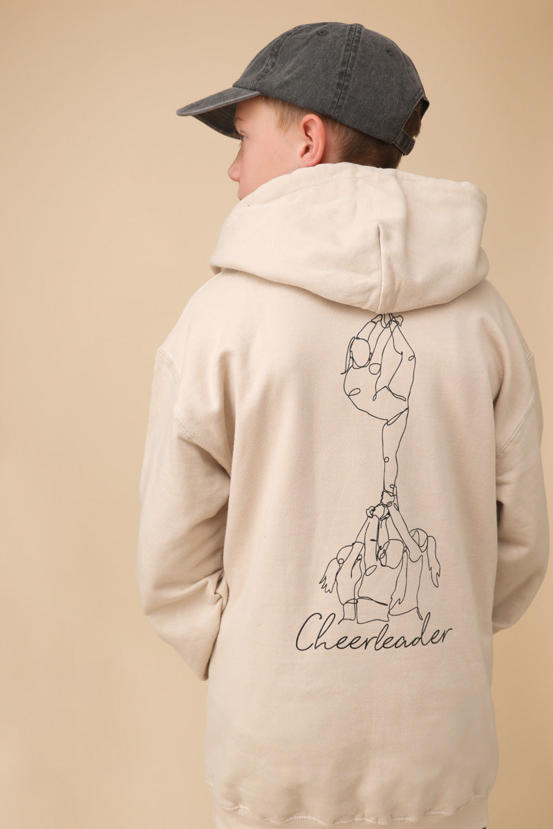 Junge steht mit dem Rücken zum Betrachter. Er trägt eine graue Capi, einen beigen Pullover mit einem großen Backprint mit einer One Line Zeichnung eines Cheerleading Groupstunts und der Aufschrift Cheerleader vor beigem Hintergrund. 