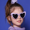 kleines Mädchen mit lila Sonnenbrille in Herzform, mit helllila Scrunchie und hell lila Pullunder steht vor blauem Hintergrund.