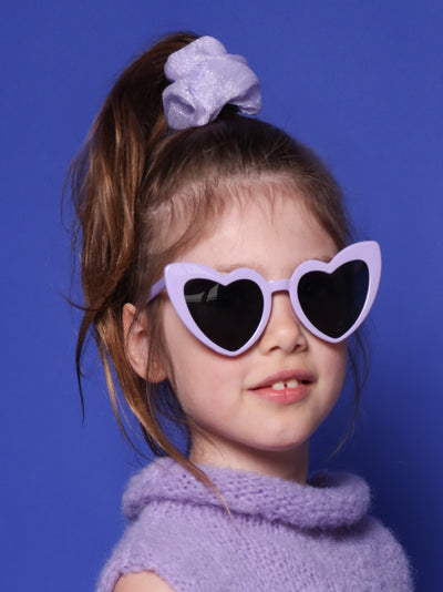 kleines Mädchen mit lila Sonnenbrille in Herzform, mit helllila Scrunchie und hell lila Pullunder steht vor blauem Hintergrund.