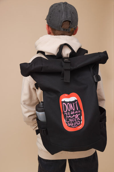 Cheer Roll Top Backpack in schwarz auf dem Rücken von einem Junior Jungen. Der Rucksack hat seitliche Gurt Justierungen und Seitentaschen für z.B. Flaschen. Auf dem Motiv ist eine weit offene Lippe mit Zähnen zu sehen. In der Öffnung vom Mund steht: Don't mess with my cheer squad please. Der Junge trägt eine Graue Jeans und graue Cap, einen Beigen Pullover und steht vor einem beigen Hintergrund. Im Rucksack ist eine Trinkflasche zu sehen. 