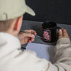 Junge sitzt an seinem Schreibtisch vor einer schwarzen Wand. Er trägt einen beigen Pullover und eine beige Cap. Er hält in der rechten Hand den "Best Cheer Friends" Becher und in der linken einen schwarzen Filzstift.