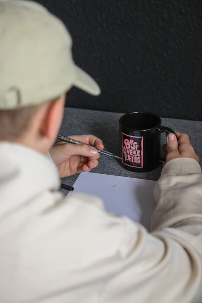 Junge sitzt an seinem Schreibtisch vor einer schwarzen Wand. Er trägt einen beigen Pullover und eine beige Cap. Er hält in der rechten Hand den "Best Cheer Friends" Becher und in der linken einen schwarzen Filzstift.