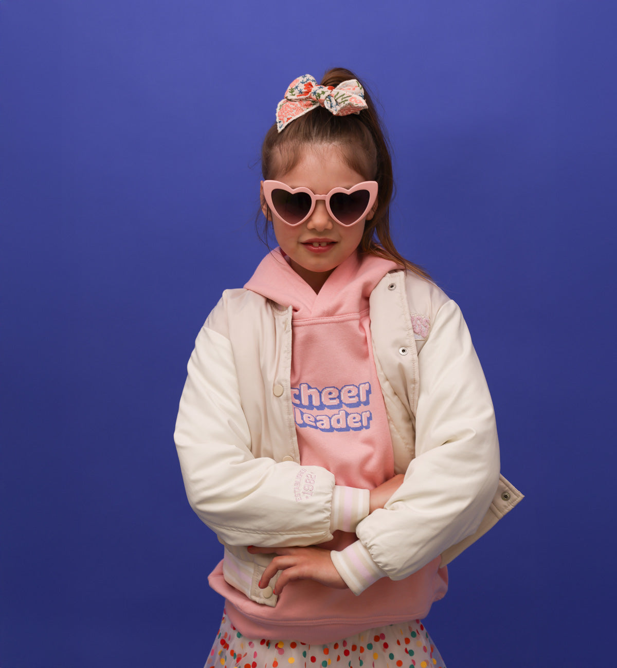 Ein kleines Mädchen mit mittelblonden Haaren, einer rosa Herzbrille, einer bestickten Schleife und einem rosafarbenem Hoodie steht vor einer blauen Wand. Der Hoode hat die Aufschrift: Cheerleader in Pixelart in lila,rosa,weiß. Sie trägt eine beige Jacke und einen bunt gepunkteten Rock. Sie ist ein Peewee. Ihre Arme sind leicht verschränkt. Sie lächelt in die Kamera.