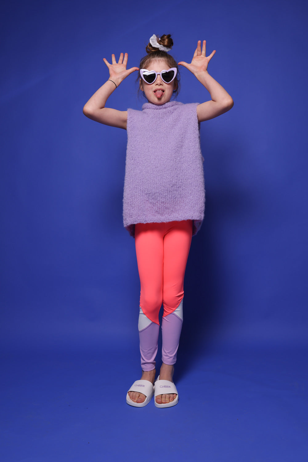 kleines Mädchen mit mittelblonden Haaren im Dutt, einer lila Herzbrille, einem hellen Scrunchie und einem lilafarbenem Wollpullunder. Sie trägt zudem eine pink-,weiß-,lilafarbene Leggings und steht vor blauem Hintergrund. Sie ist ein Peewee. Ihre Arme sind in Jazzhands an ihrem Kopf. SIe streckt die Zunge raus. An ihren Füßen sind weiße Badelatschen mit der Aufschrift: Cheer in Lila.