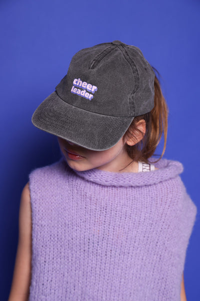 Zu sehen ist ein Cheerleader Pixelart Motiv auf einer Relaxed Vintage Cap in der Farbe vintage black ist hier auf dem Kopf von einem kleinen Mädchen zu sehen. Das Mädchen trägt einen lilafarbenen Pullunder und steht vor einem blauen Hintergrund. Das Gesicht ist bis auf die Lippen von der Cap versteckt.