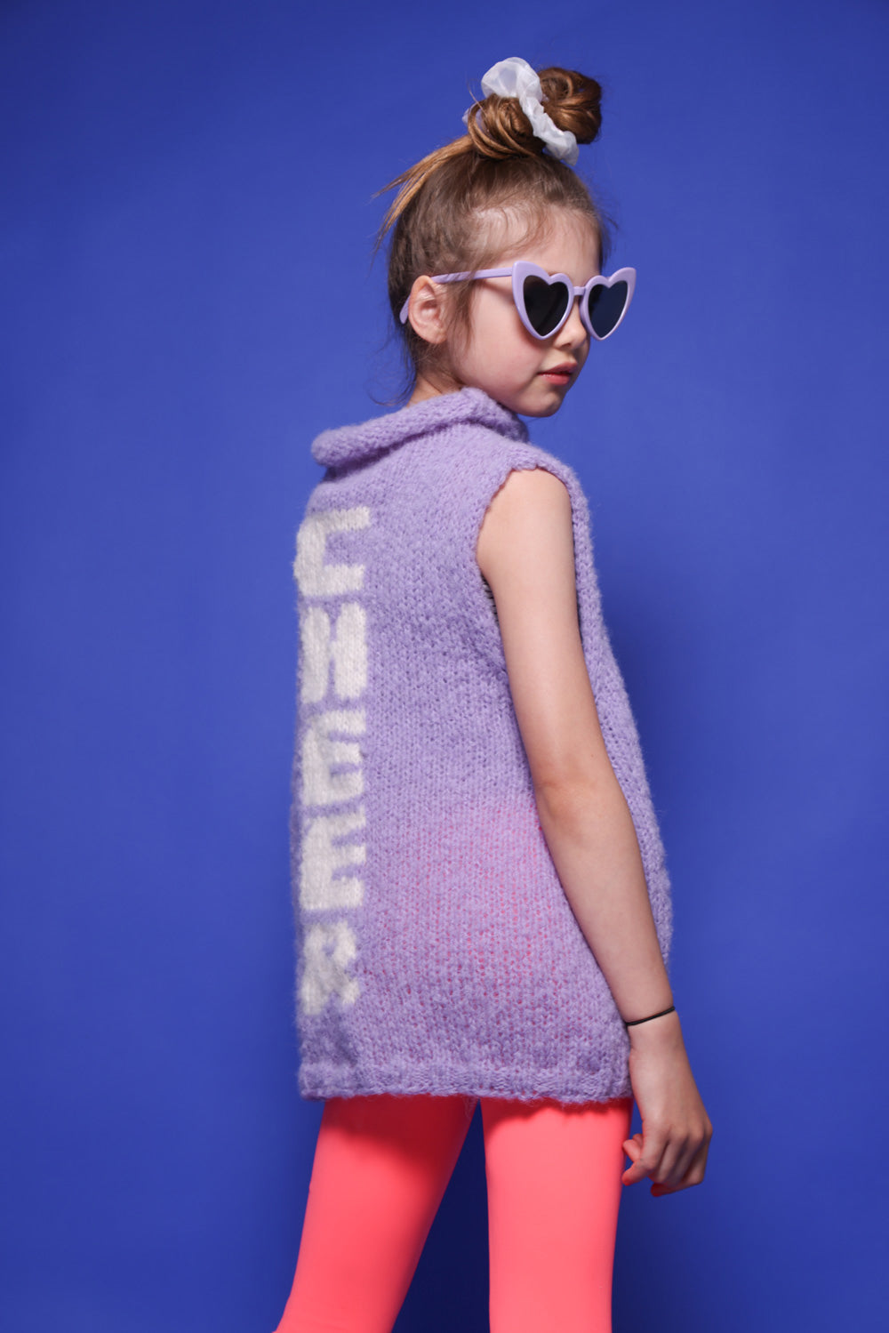 Ein kleines Mädchen mit mittelblonden Haaren im Dutt, einer lila Herzbrille, einem hellen Scrunchie und einem lilafarbenem Wollpullunder steht vor einer blauen Wand. Sie trägt zudem eine pink-,weiß-,lilafarbene Leggings. Sie ist ein Peewee. Auf der Rückseite des Pullunders ist das Wort Cheer eingestrickt. Die Buchstaben laufen untereinander. 