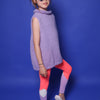 kleines Mädchen mit mittelblonden Haaren im hohen Cheerzopf, einer lila Herzbrille, einem lila Scrunchie und einem lilafarbenem Wollpullunder. Sie trägt zudem eine pink-,weiß-,lilafarbene Leggings und steht vor blauem Hintergrund. Sie steht seitlich und hat ein Bein angewinkelt. Sie ist ein Peewee und lächelt. An ihren Füßen sind weiße Badelatschen mit der Aufschrift: Cheer in Lila.
