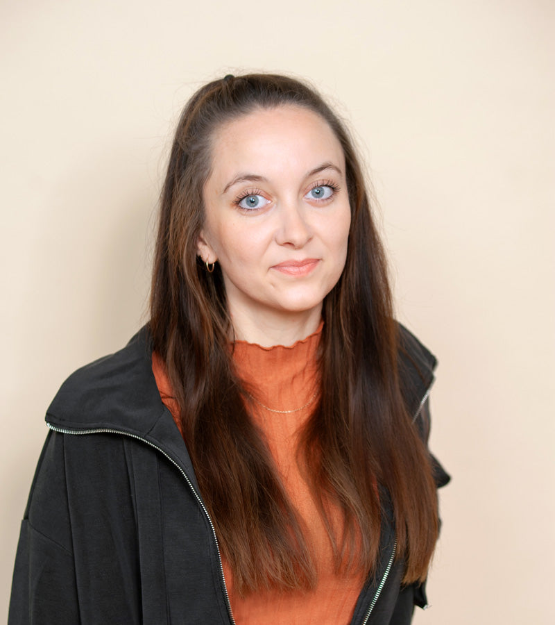 Zuza, die Frau hinter dem Bow Bow shop steht vor einer beigen Wand. Sie trägt ein bronzefarbenes Oberteil und eine dunkel-graue Jacke und hat blau-grüne Augen und dunkle braune Haare.